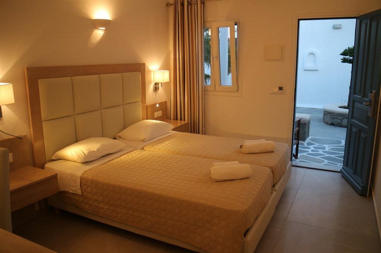A hotel room with two beds, a wooden headboard, bedside lamps, and a view of the courtyard through an open door.