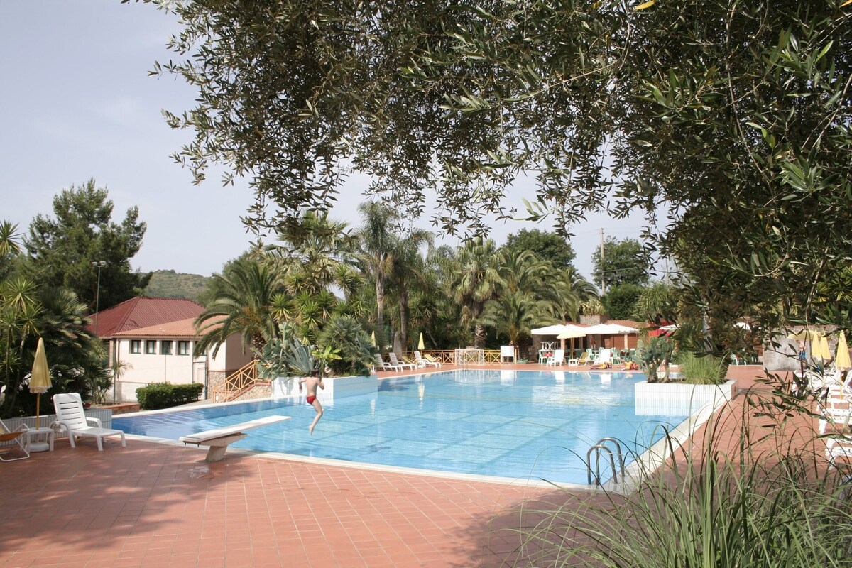 Une piscine entourée de chaises longues et de parasols, bordée de palmiers et d’un bâtiment au toit rouge.