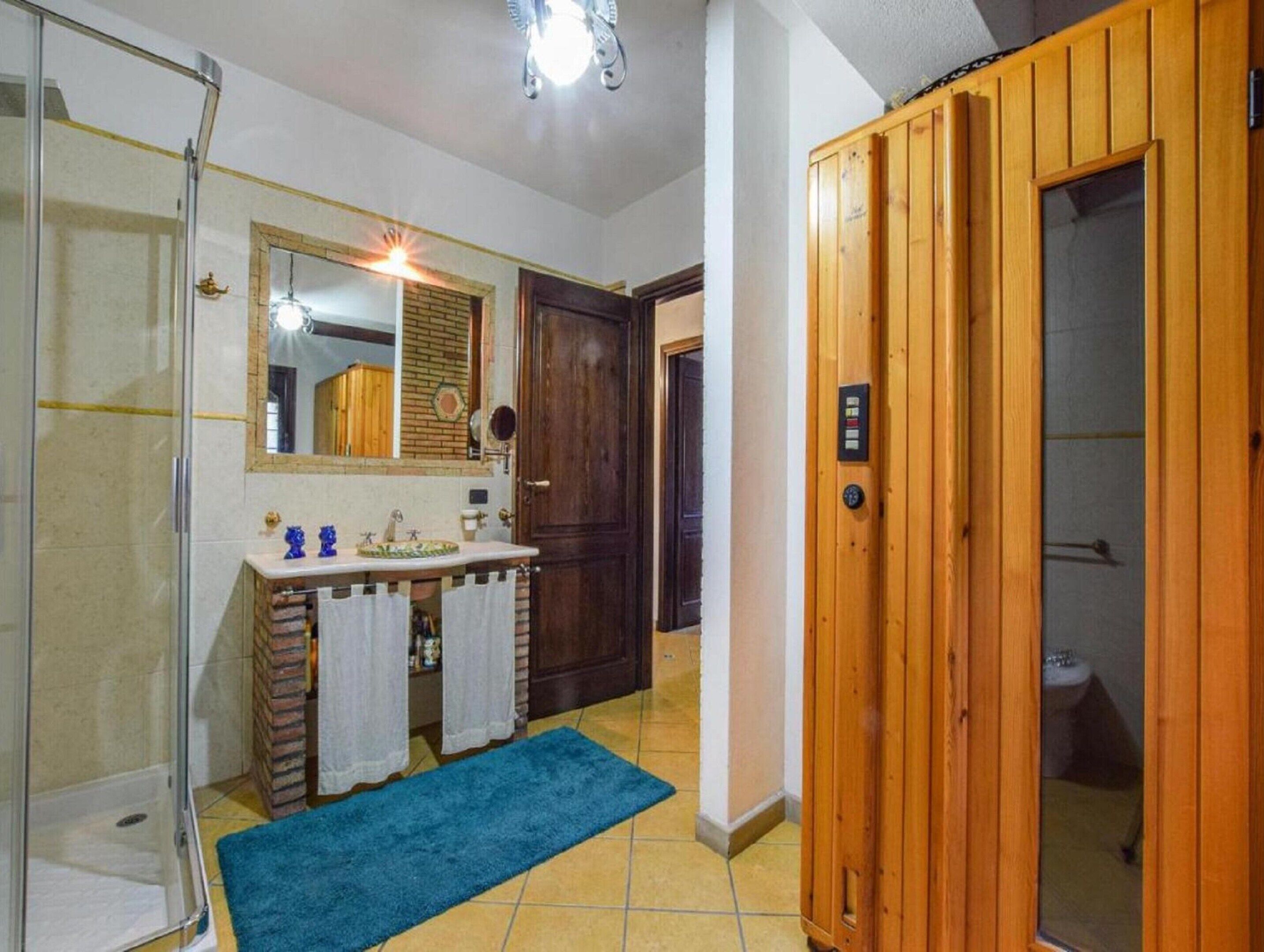 A bathroom with a glass shower enclosure, a wooden door, a sink with a mirror, and a blue bath mat.