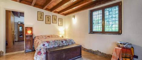 Ein Schlafzimmer mit Holzbalkendecke, einem Bett mit floralem Überwurf, einer Bank, einem Fenster mit Aussicht und gerahmten Bildern an der Wand.