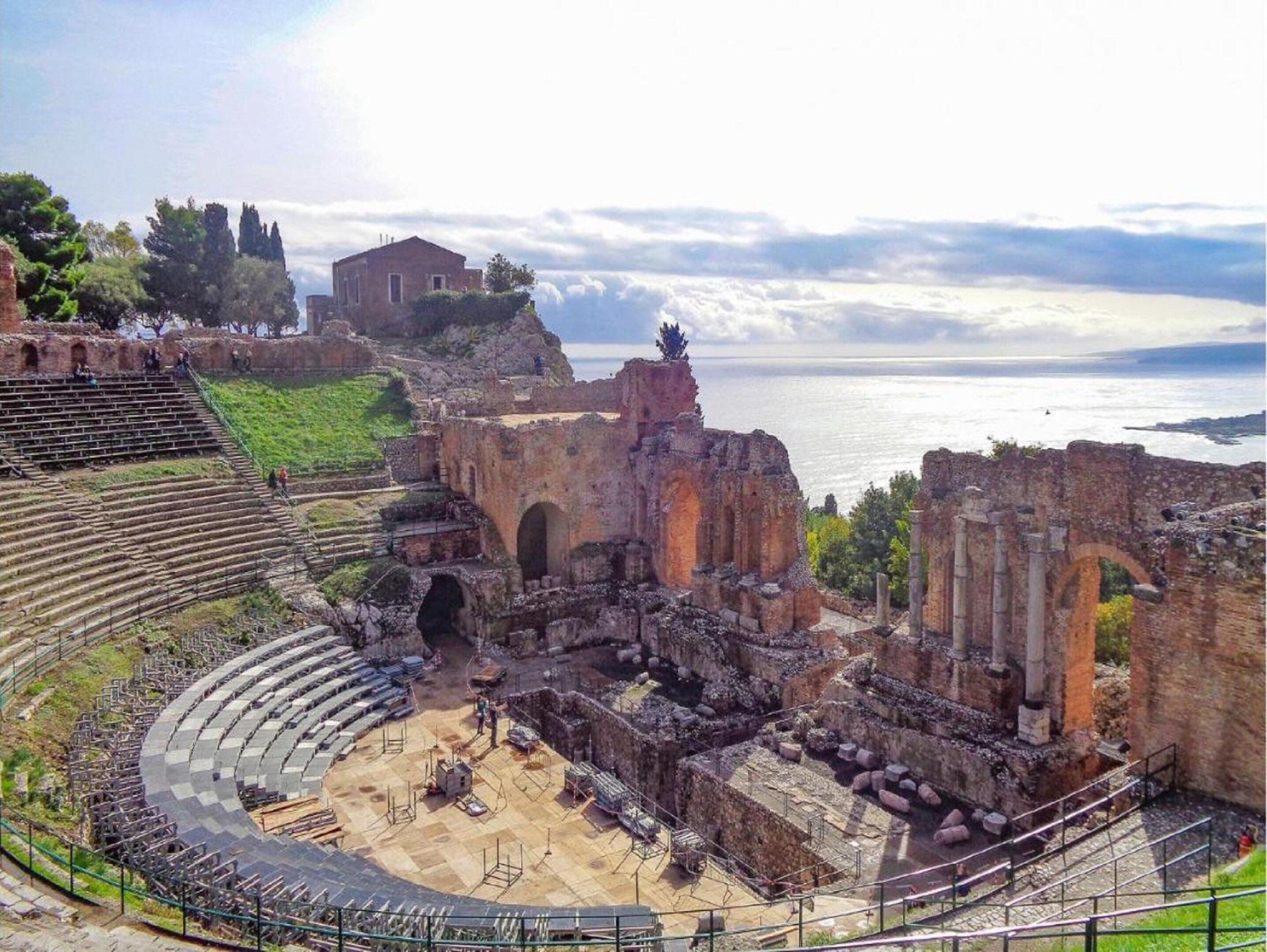 Un ancien amphithéâtre dont la scène et les gradins sont partiellement conservés, entouré de ruines et dominant une étendue d’eau.