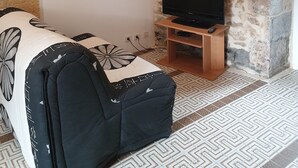 A living room with a stone wall, a flat-screen TV on a wooden stand, a sofa with a patterned cover, and a patterned floor. - Studio 2 - 3 min from Futuroscope - 1 to 4 people (Chasseneuil-du-Poitou)