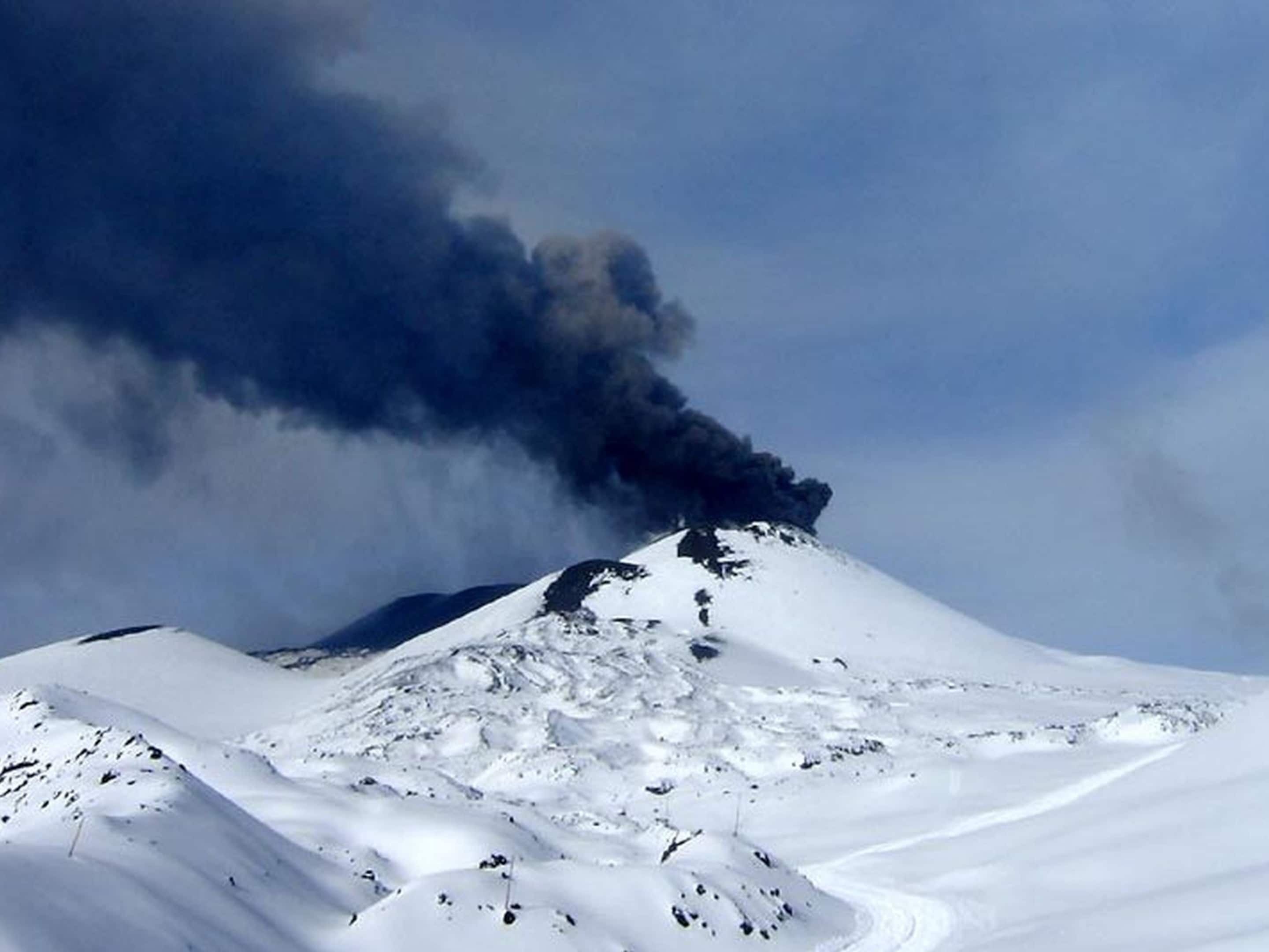 Uma montanha coberta de neve com uma coluna de fumaça escura a subir do cume.