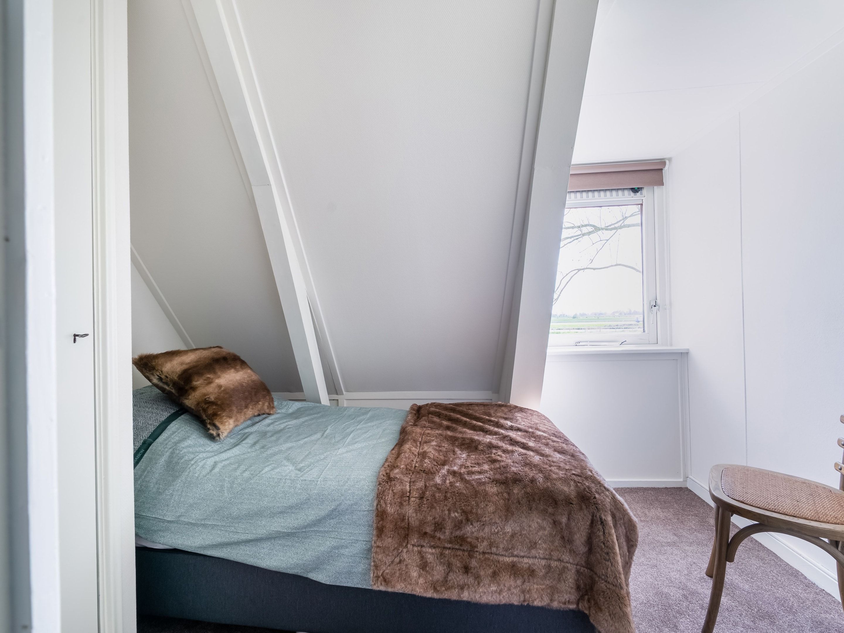 A bedroom with a sloped ceiling, a bed with a blue and brown blanket, a window with a view of the outdoors, and a wooden chair.