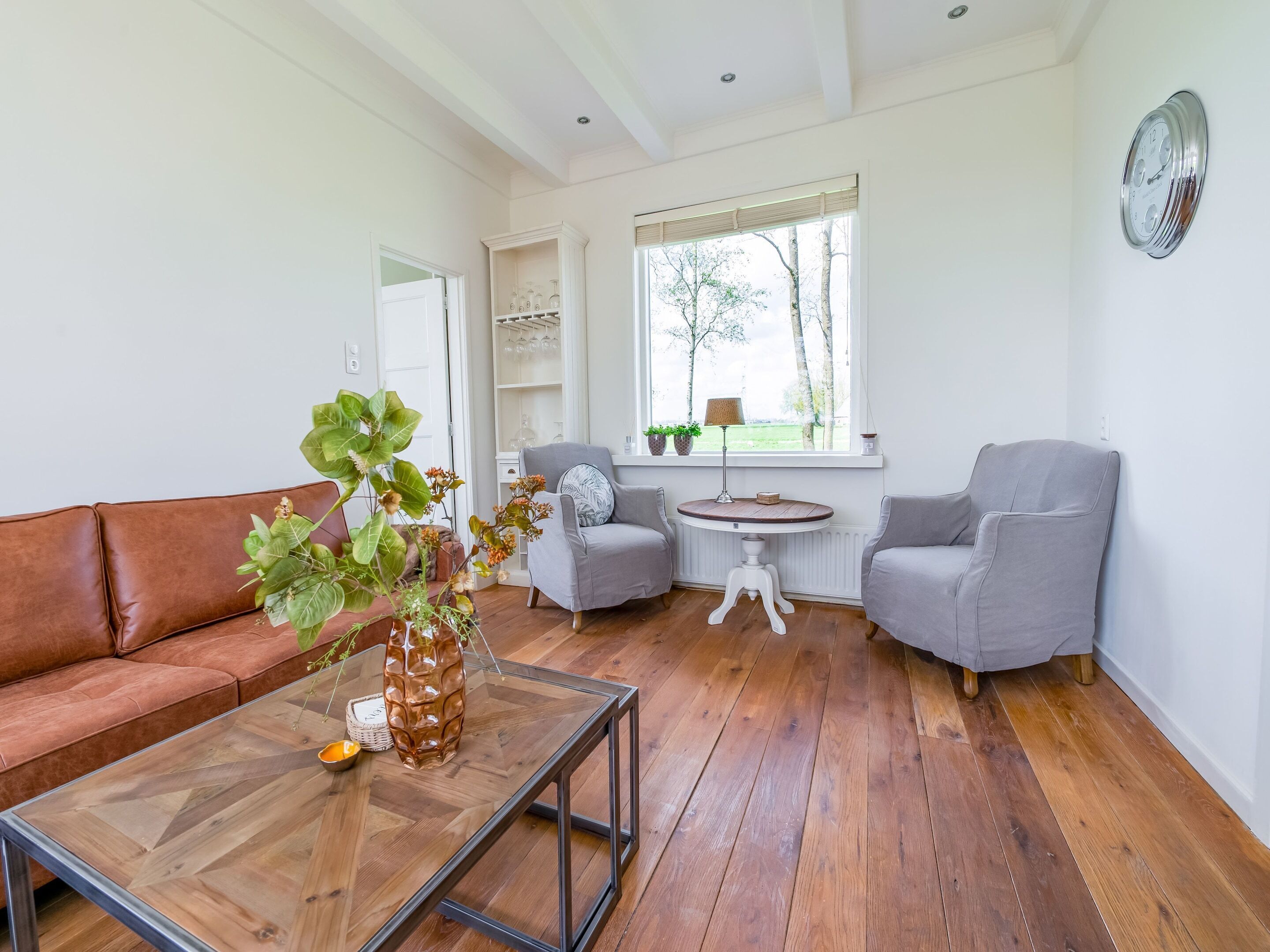 A living room with a brown leather sofa, a glass coffee table, two grey armchairs, a round wooden table, and a window with a view of trees.