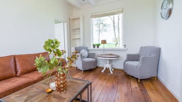 A living room with a brown leather sofa, a glass coffee table, two grey armchairs, a round wooden table, and a window with a view of trees.