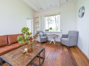 A living room with a brown leather sofa, a glass coffee table, two grey armchairs, a round wooden table, and a window with a view of trees. - B&B Farm Grutsk in Suwald With Private Harbour (Suwâld)