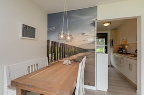A dining area with a wooden table, two hanging light fixtures, a wall-mounted picture, and a view into the kitchen.