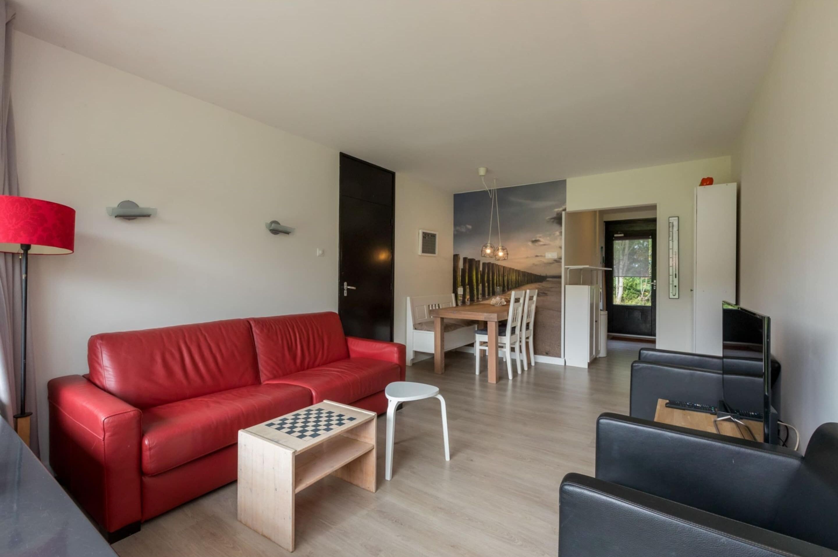 A modern living room with a red sofa, a small wooden table with a chessboard, a white chair, a dining area with a wooden table and chairs, a TV, and a staircase leading to another level.