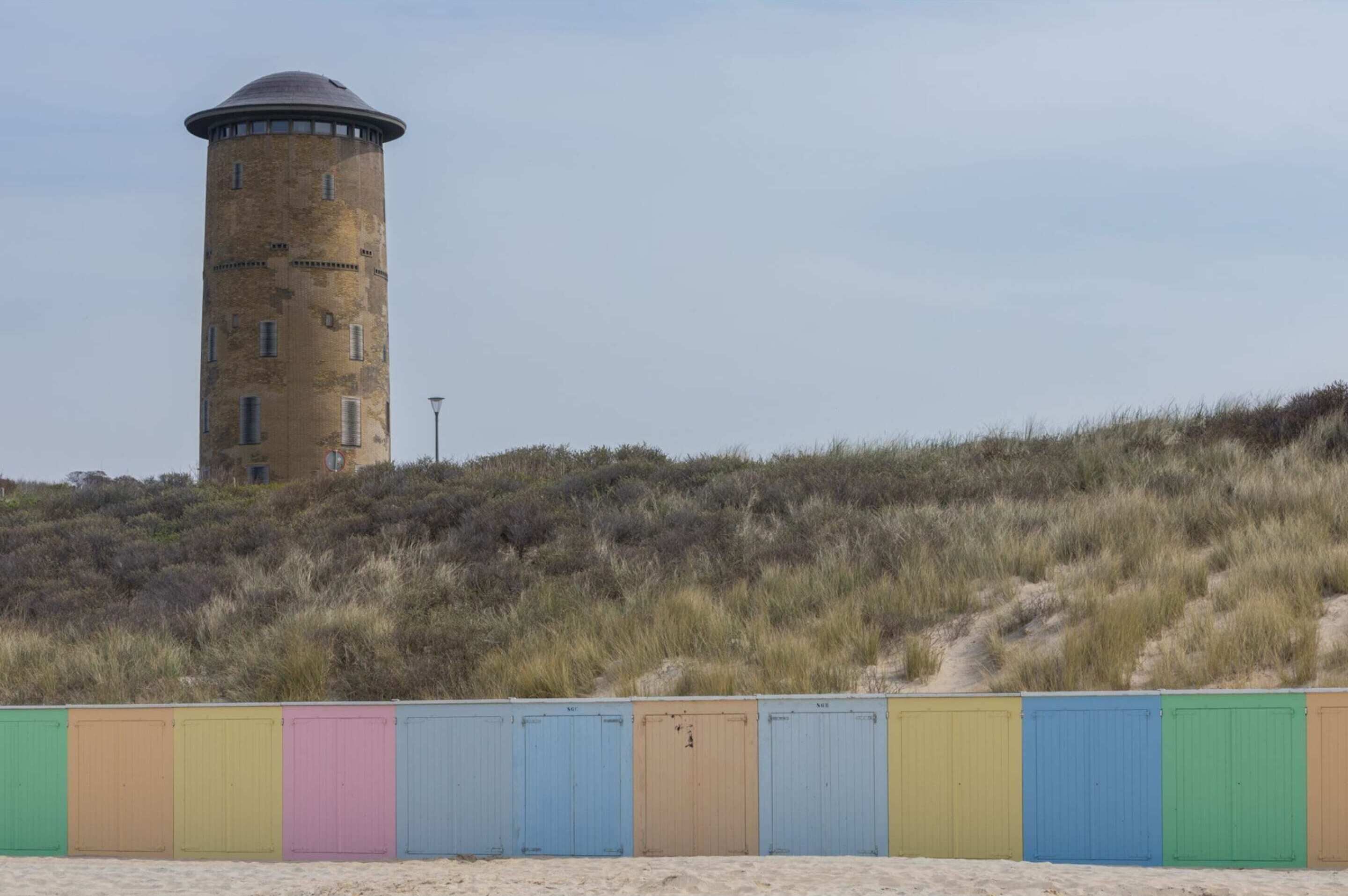 A cylindrical water tower on a grassy hill with a row of colorful beach huts in the foreground.