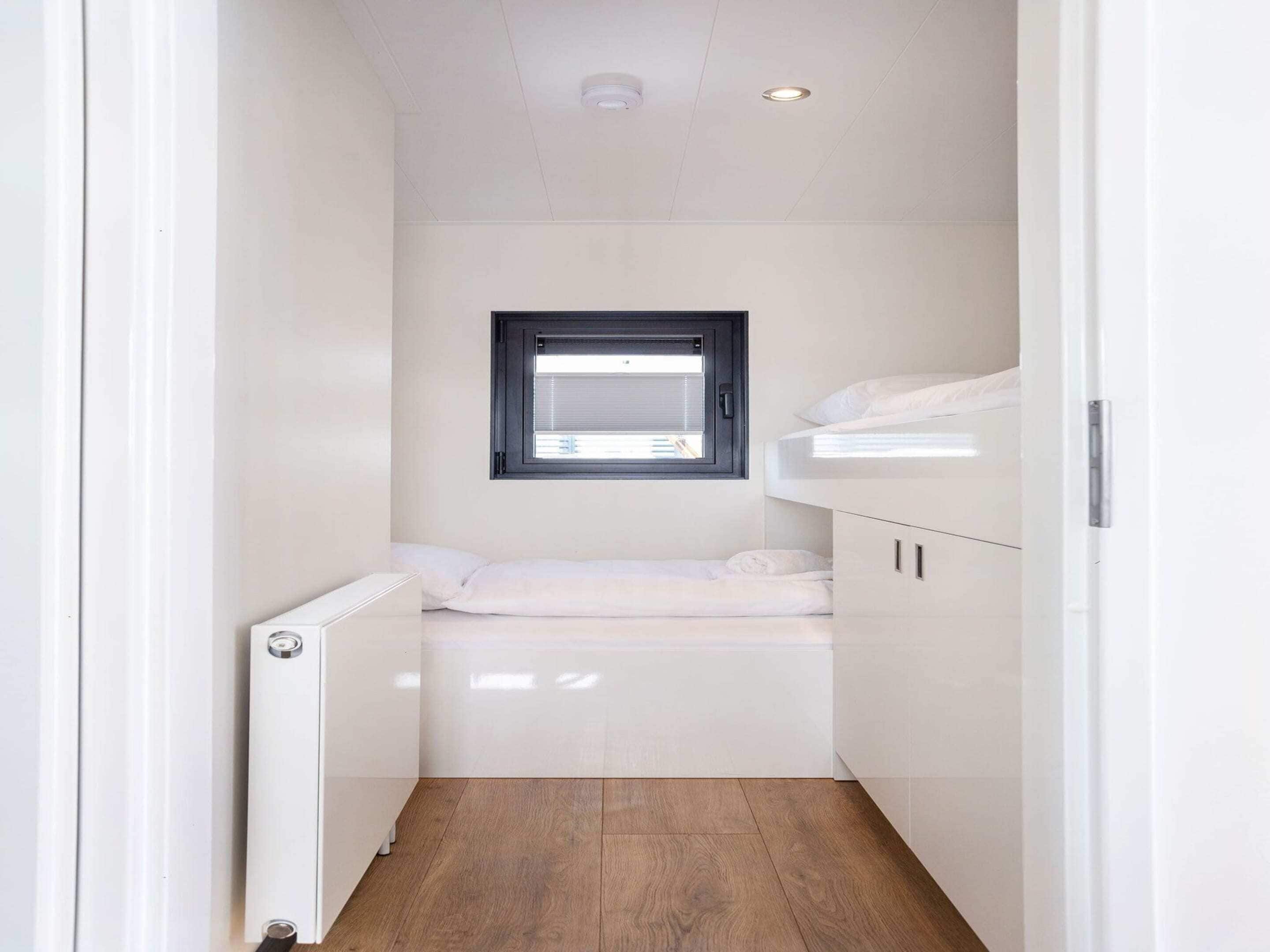 A narrow, white-walled room with a small window, a bunk bed, and a radiator.