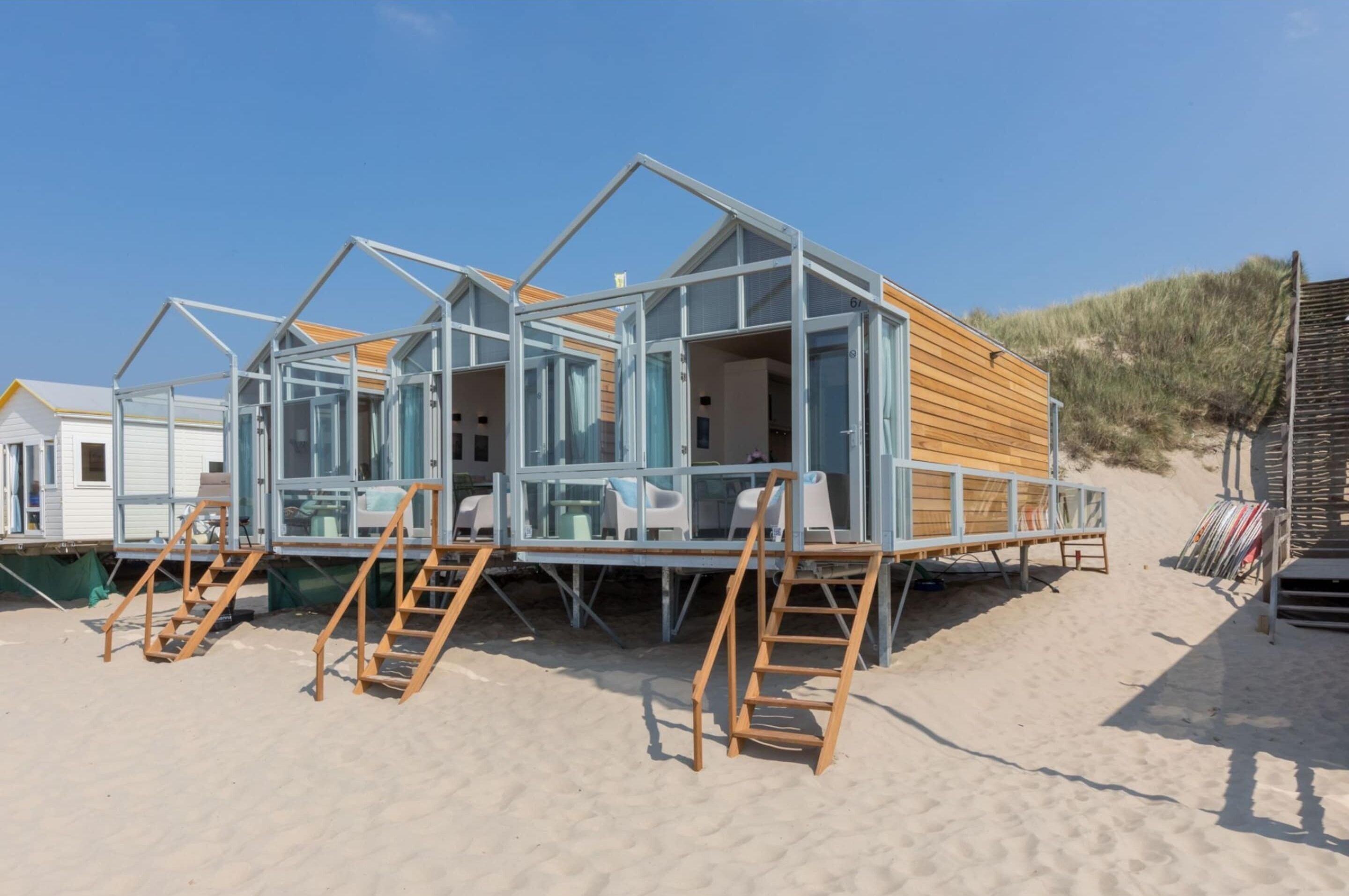 A beach house with a wooden deck, glass walls, and a sandy beach setting.