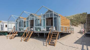 A beach house with a wooden deck, glass walls, and a sandy beach setting.