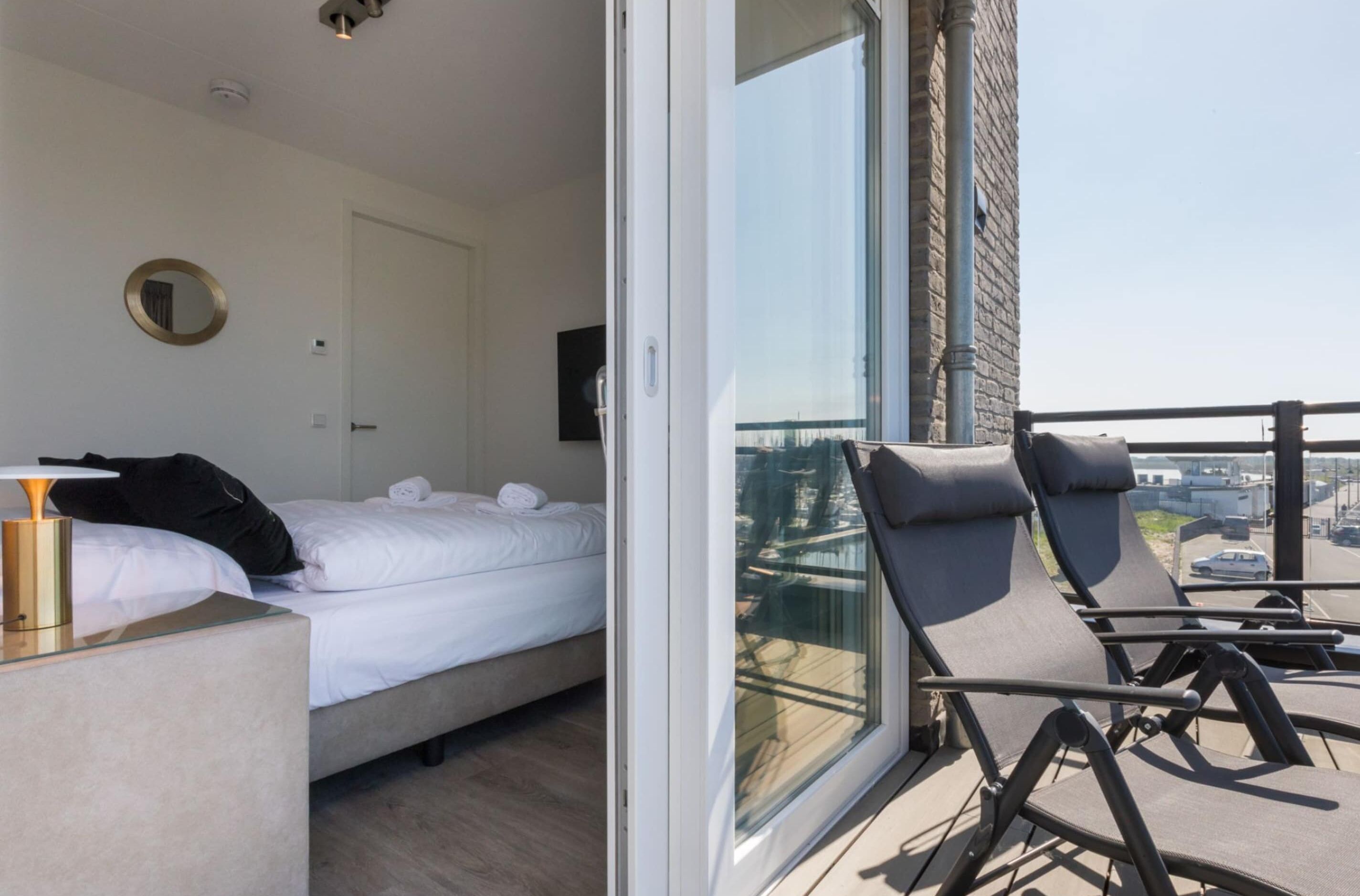 Habitación de hotel con cama, mesita de noche, una silla y un balcón con vistas a la ciudad.
