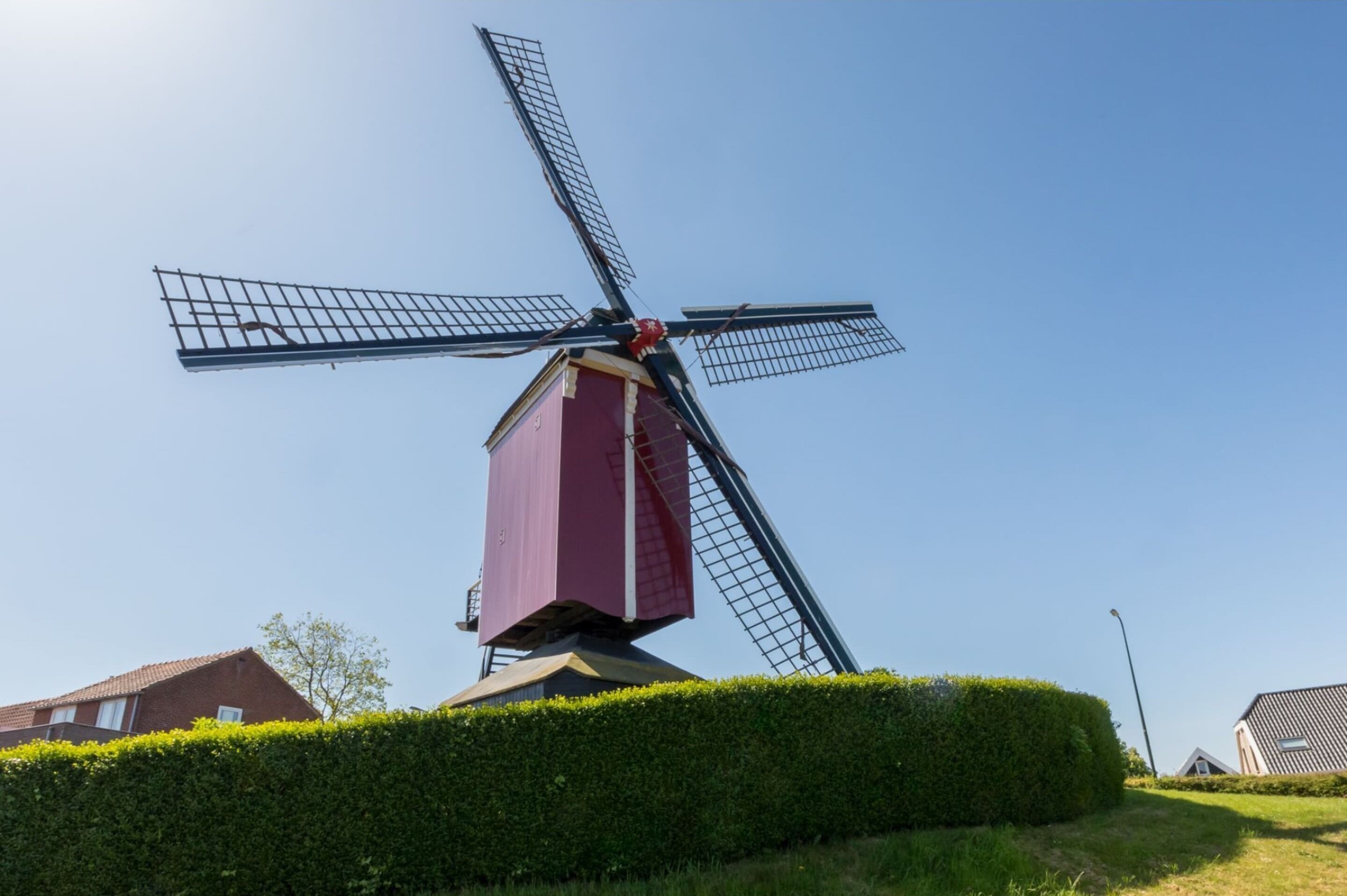 A windmill with a red base and a metal structure, surrounded by a well-maintained hedge and a clear blue sky.