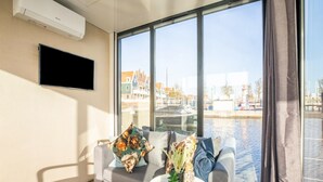 A modern living room with a grey sofa, a small white table, and a large window offering a view of a canal and buildings. - Beautiful Houseboat With Shared Pool (Volendam)