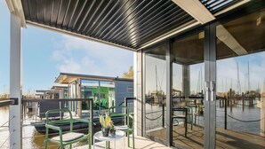 A modern outdoor deck with a glass enclosure, green chairs, and a table with a plant. - Beautiful Houseboat With Shared Pool (Volendam)
