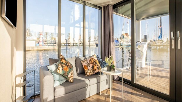A modern living room with a sofa, a small table, and a view of a marina through large windows. - Beautiful Houseboat With Shared Pool (Volendam)