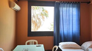 A room with a bed, a table with a tablecloth, a chair, a window with curtains, and an air conditioning unit.