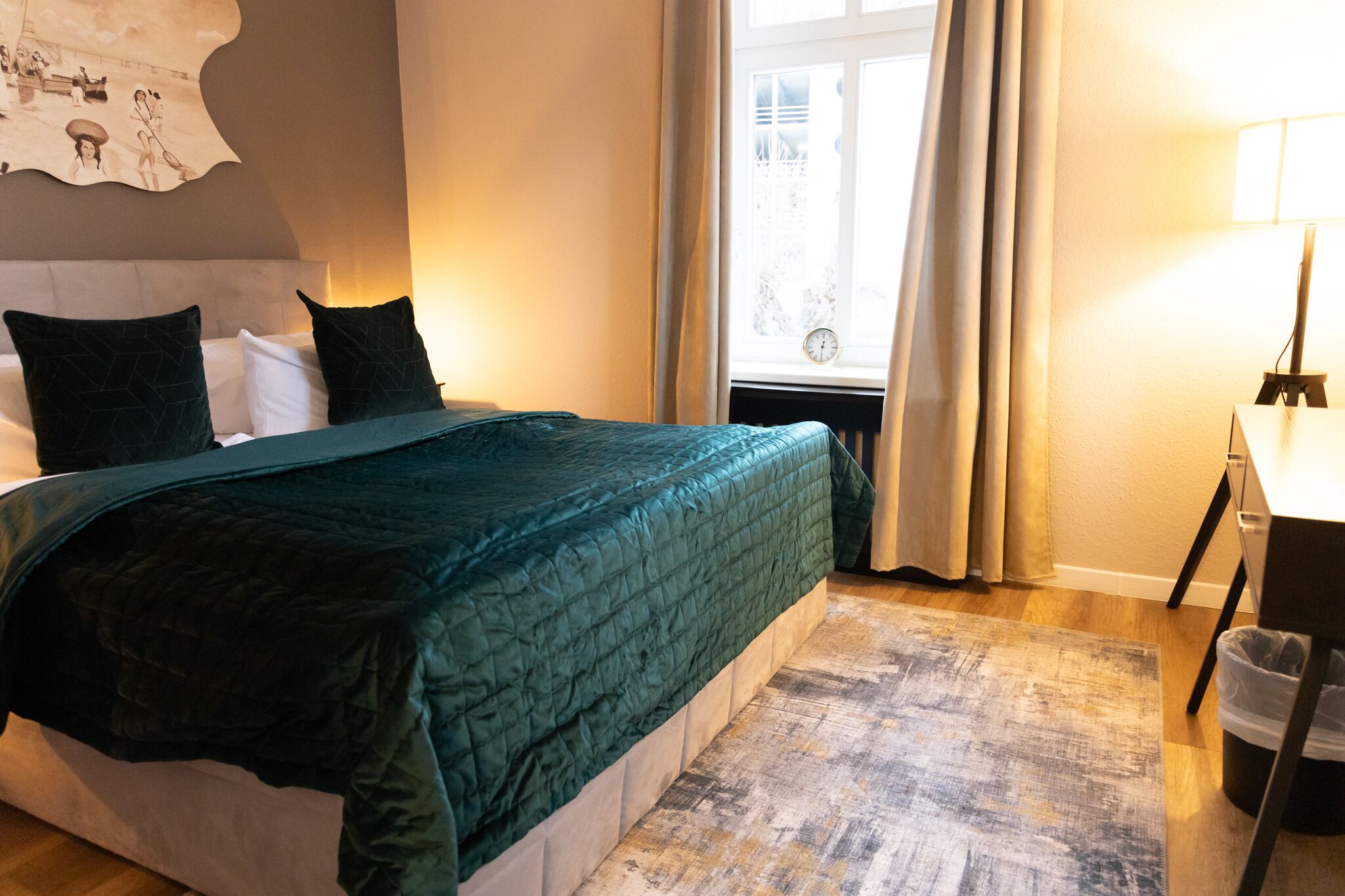 Un letto rifatto con un copriletto verde scuro, una testiera e due cuscini neri. Si vede una finestra con tende e una lampada su un comodino.