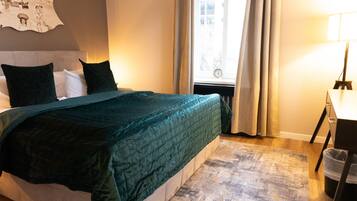 Ein ordentlich bezogenes Bett mit einer dunkelgrünen Tagesdecke, einem Kopfteil und zwei schwarzen Kissen. Ein Fenster mit Vorhängen ist sichtbar, und eine Lampe steht auf einem Nachttisch.