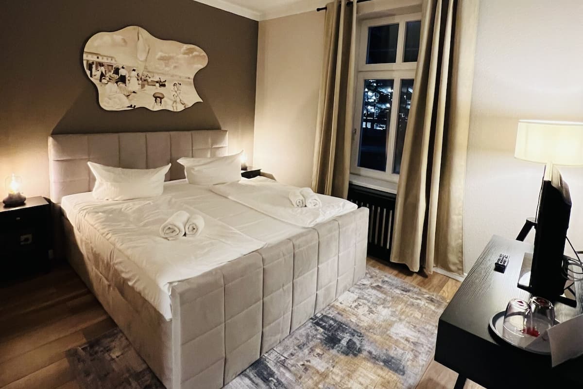 A bedroom with a large bed, a headboard with a decorative panel, a bedside table with a lamp, a window with curtains, and a desk with a computer.