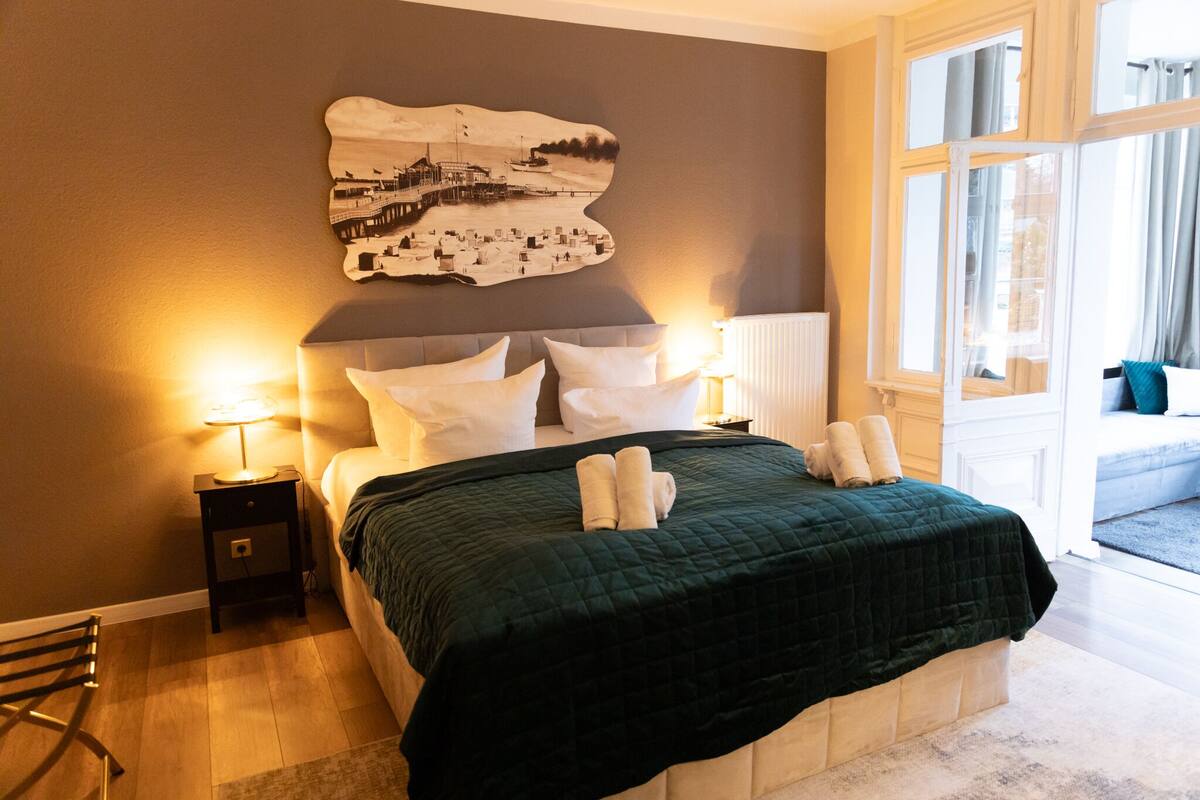 A neatly made bed with a green quilt, two bedside tables with lamps, a large wall-mounted picture, and a view of the outdoors through a window.