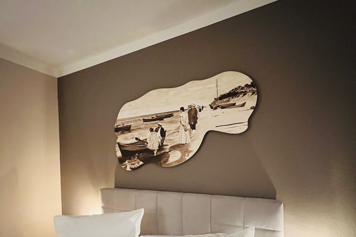 A hotel room with a bed, a headboard, a nightstand with a lamp, and a wall-mounted artwork.