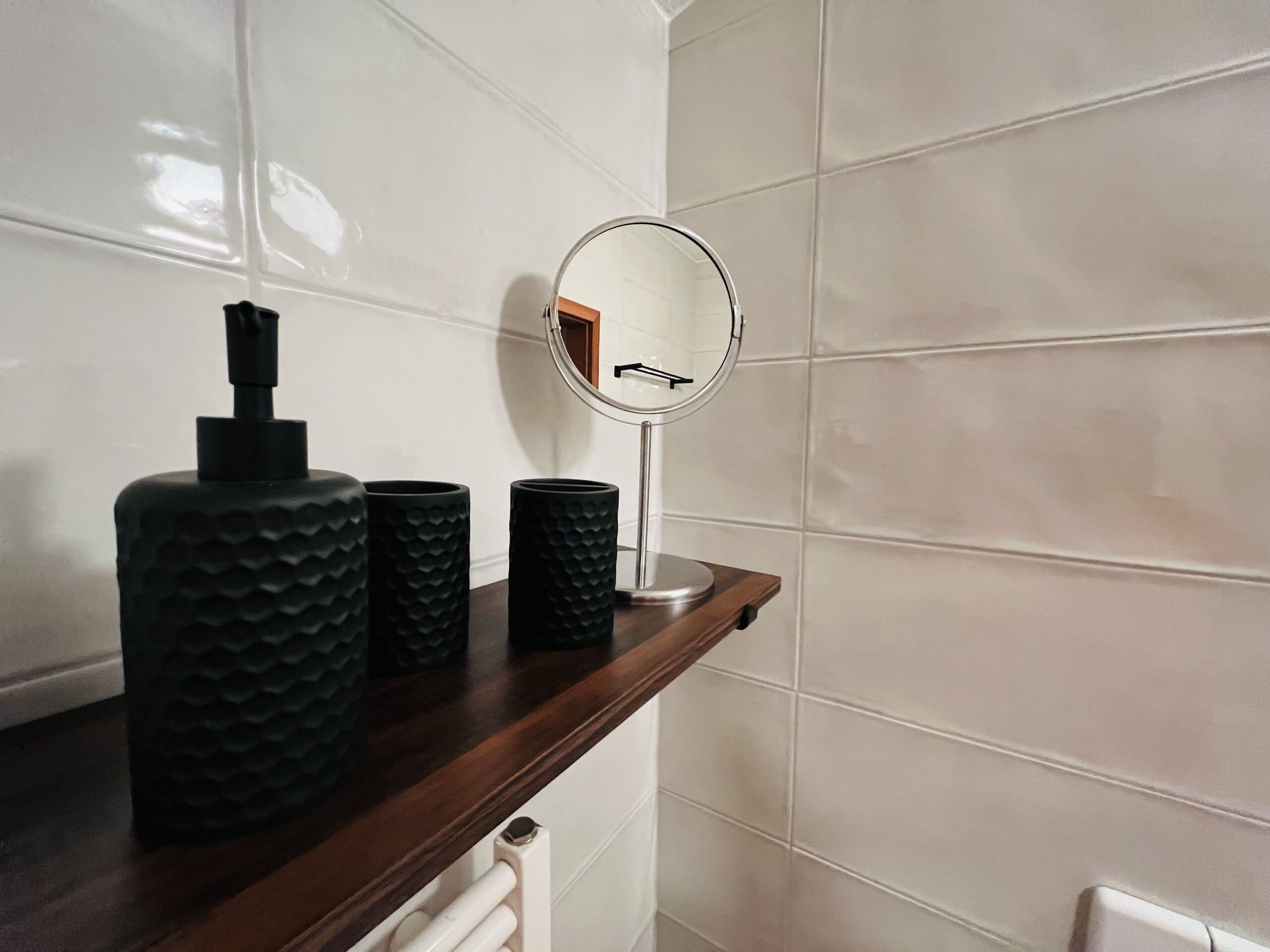 Un baño con baldosas blancas, un estante de madera con tres recipientes negros con textura, y un espejo redondo con marco de madera.