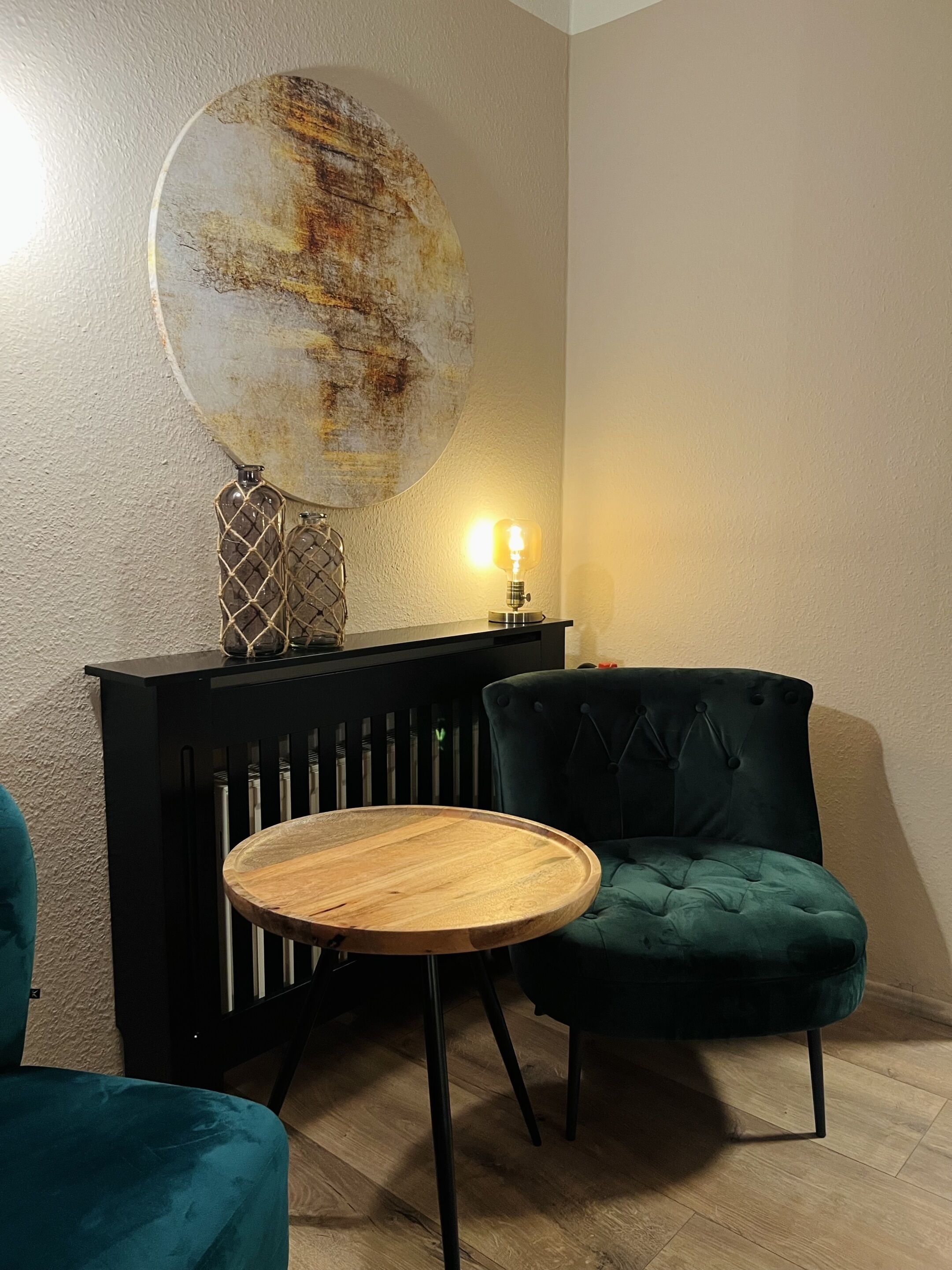 Una habitación con una mesa redonda de madera, una silla tapizada en verde oscuro, un aparador negro con una lámpara decorativa y un cuadro abstracto grande en la pared.