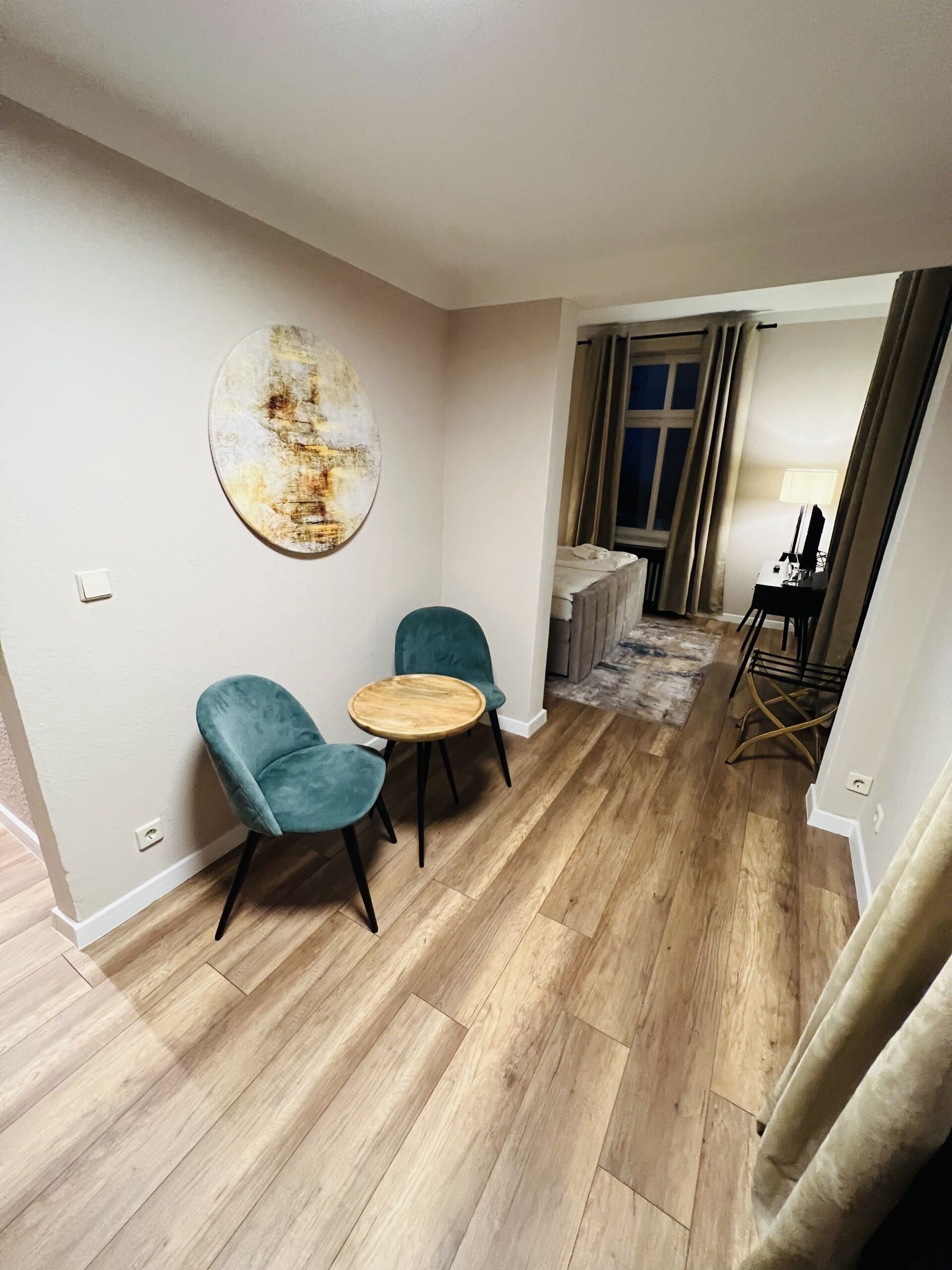 A room with a wooden floor, two teal chairs, a round wooden table, a large abstract painting, and a bed in the background.