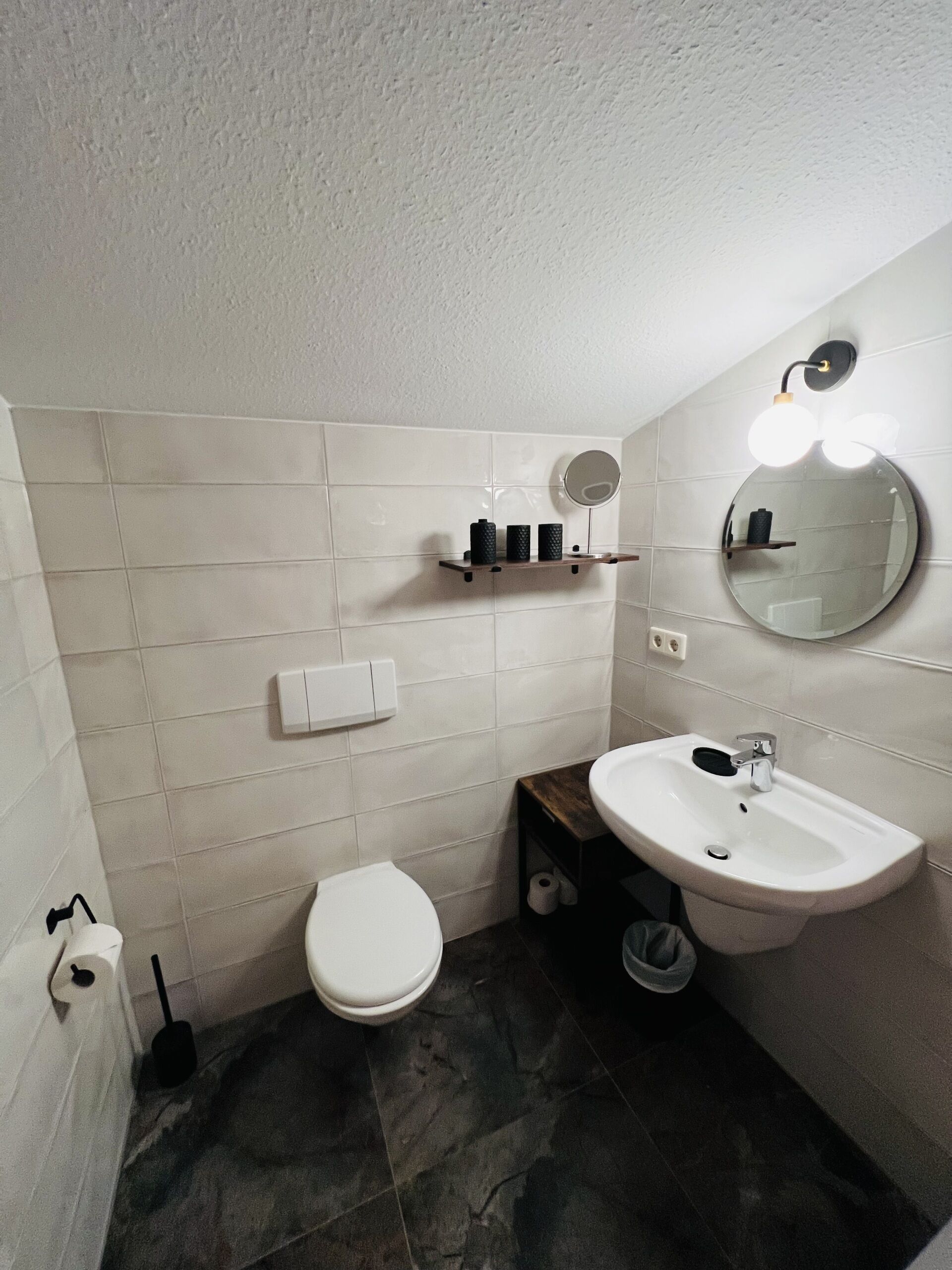 A bathroom with white tiles, a white toilet, a sink with a mirror, and a shelf with toiletries.