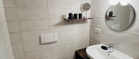 A bathroom with white tiles, a white toilet, a sink with a mirror, and a shelf with toiletries.