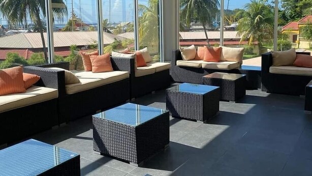 A sunlit lounge area with large windows, wicker furniture, and a view of palm trees and buildings. - West Palm Hotel (Chaguaramas)