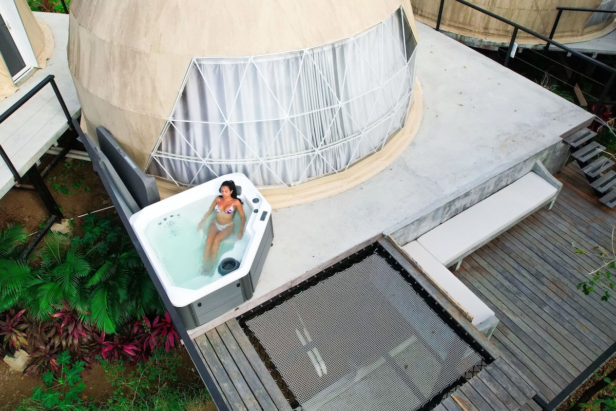 A hot tub with a person sitting in it, surrounded by a concrete deck and a covered seating area.