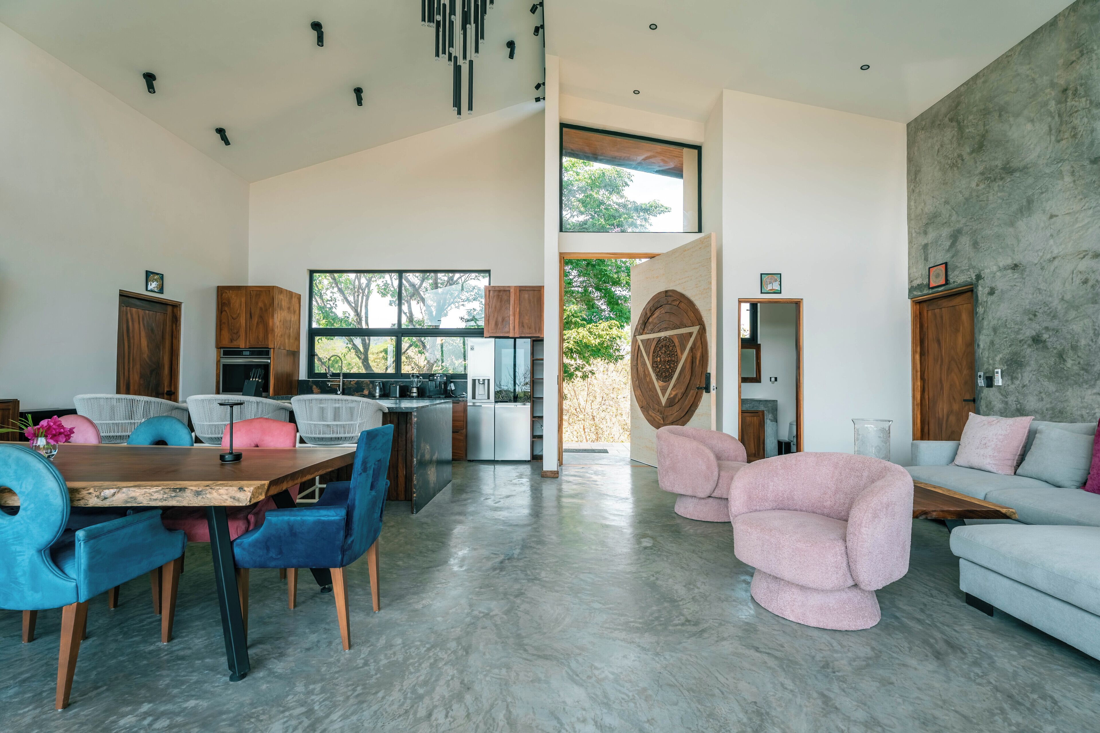 A spacious living area with a dining table, chairs, and a sofa. There is a kitchen area with cabinets and a countertop. The room has large windows and a view of trees outside.
