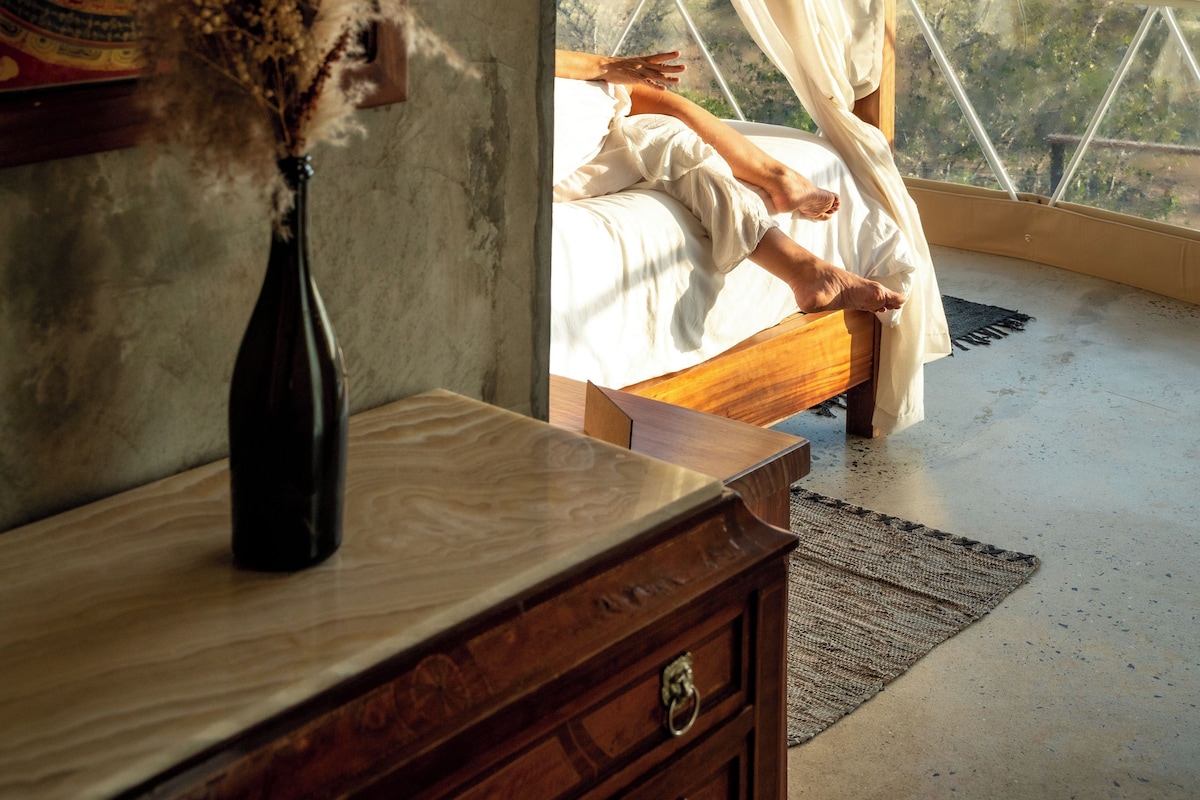 Um quarto com uma janela grande, uma cama, uma cômoda de madeira, um vaso com flores secas e uma tapeçaria colorida na parede.