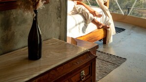 Un dormitorio con un ventanal grande, una cama, un aparador de madera, un florero con flores secas y un tapiz colorido en la pared.