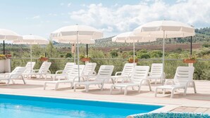 A pool area with white lounge chairs and umbrellas, a tiled deck, and a view of hills in the background.