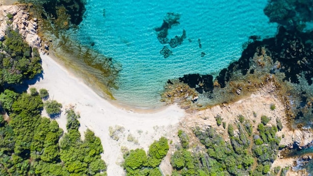 An aerial view of a coastal area with clear turquoise waters, sandy beach, and dense green vegetation.