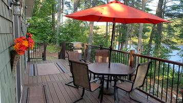 A wooden deck with a table and chairs, a red umbrella, and a view of a lake surrounded by trees.
