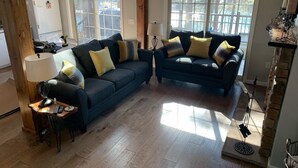 A living room with two dark blue sofas, a wooden coffee table, and a lamp. There are windows with a view of trees and a white refrigerator in the background.