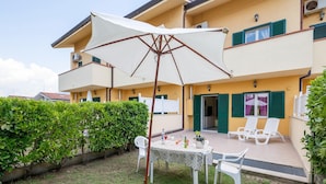 A courtyard with a table and chairs, a white umbrella, and a building with green shutters. - Beachside Villa in Paestum (Capaccio-Paestum)