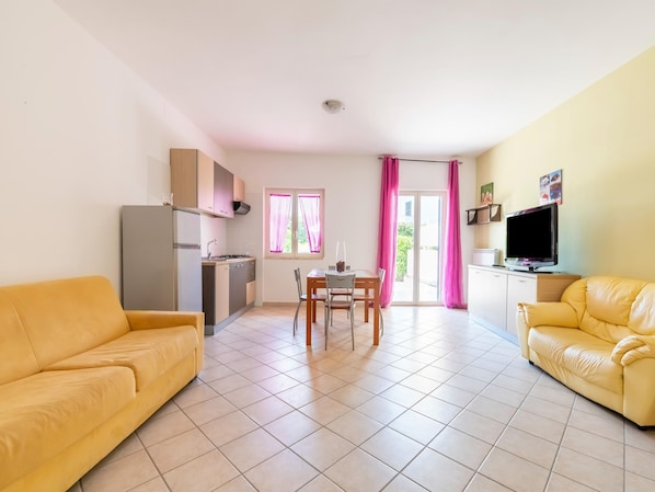 A living room with a sofa, a dining table with chairs, a kitchen area, and a TV.