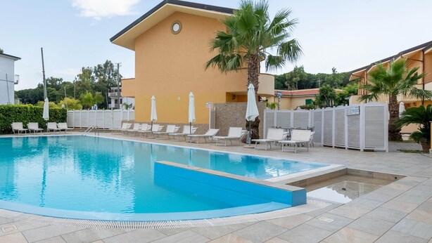 A swimming pool with lounge chairs and umbrellas, surrounded by a fence and palm trees. - Beachside Villa in Paestum (Capaccio-Paestum)