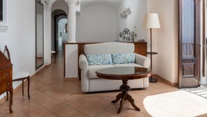 A living room with a sofa, a round table, and a lamp. There is a wooden chair and a cabinet. The room has a tiled floor and white walls with arches.