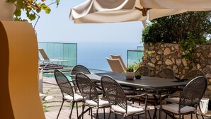 An outdoor patio with a table and chairs, a large umbrella, and a view of the sea.