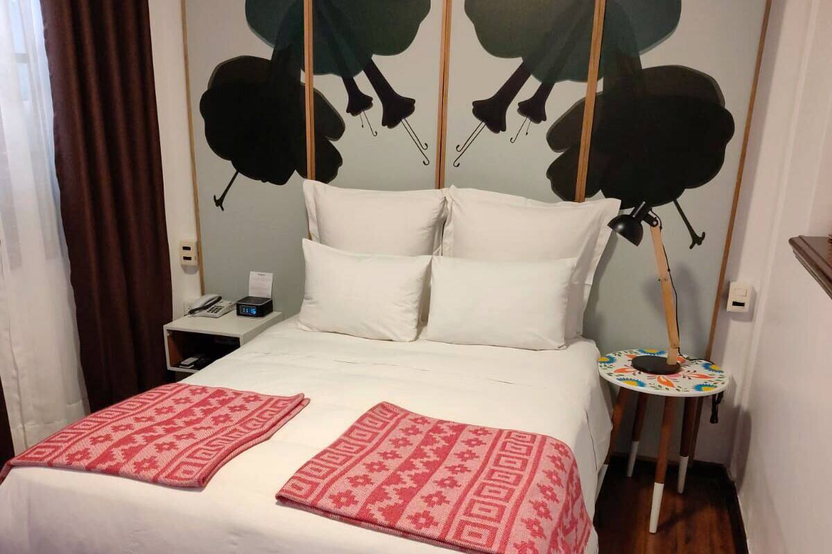 Una cama bien hecha con sábanas blancas y almohadones estampados rojos, una mesita de noche con lámpara, y una obra de arte en la pared sobre la cama.