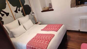 Un dormitorio con cama, almohadas, mesita de noche con lámpara, estante pequeño con adornos y un radiador.