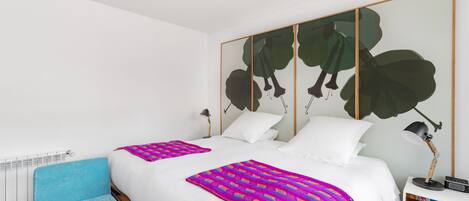 A modern hotel room with a large bed, a blue armchair, a colorful blanket, and a large abstract painting on the wall.
